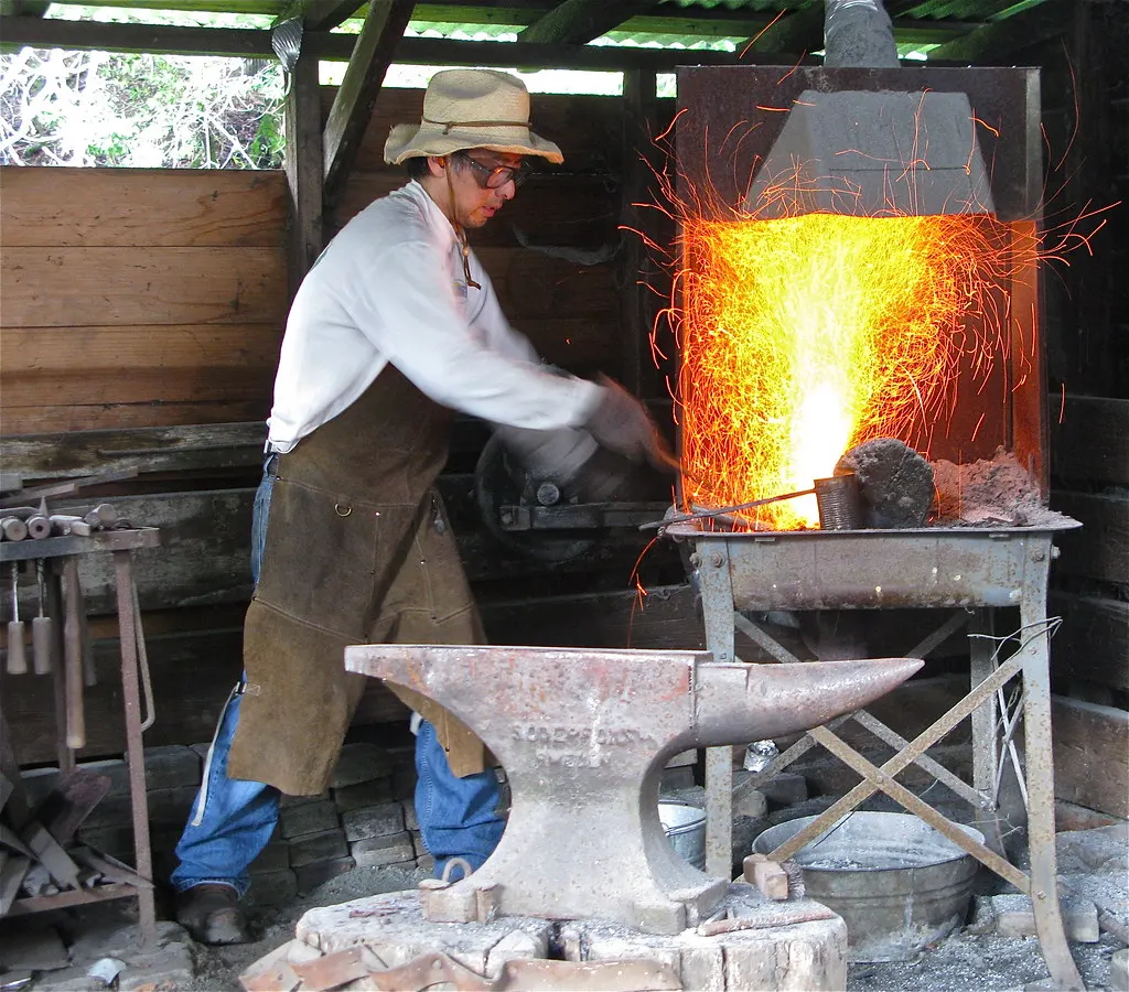 Herrero que trabaja junto al yunque mientras el fuelle aviva la forja
